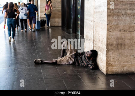 Rome, Italy - September 4, 2018: African refugee homeless man lying on sidewalk ground in city by entrance to Termini rail station, people walking Stock Photo