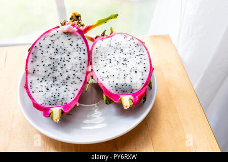 Closeup of cut open pink purple dragon dragonfruit fruit slices halves half inside white flesh, seeds cactus on plate, wooden table by window Stock Photo