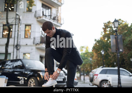 Attractive man stopped on the street to bind laces on his white sneakers. Young fashionable businessman. Stock Photo