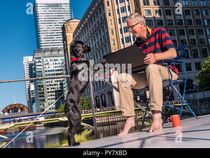 Man & dog happily networking on deck of his river barge home office enjoying sun & his pet spaniel, big business city buildings Canary Wharf London Stock Photo