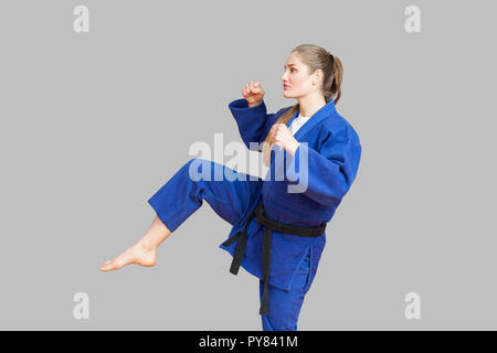 Side view of agressive athletic karate woman in blue kimono with black belt are ready to fight, punching with foot and looking away. Japanese martial 