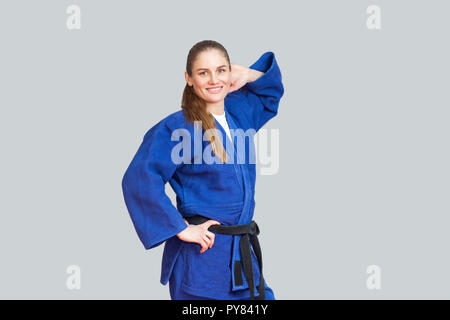 Beautiful happy athletic karate woman in blue kimono with black belt posing and holding hand behind her head are looking at camera. Japanese martial a Stock Photo