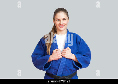 Portrait of happy beautiful athletic karate woman in blue kimono with black belt standing and looking at camera with toothy smile. Japanese martial ar