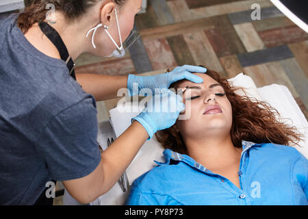 microblading, specialist drawing eyebrow shape with pencil. woman laying on bed, eyes closed. Stock Photo