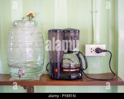 Cooler bottle and electric kettle with plug on wooden shelf on light green container wall background in cafe. Stock Photo