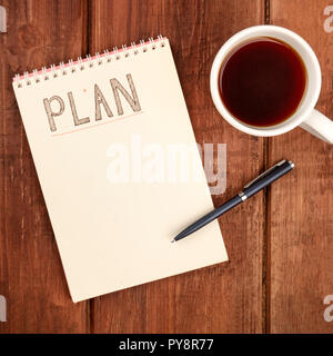 A photo of a spiral notepad with the word Plan, with a cup of coffee and a pen, shot from above on a dark rustic wooden desk with copy space Stock Photo
