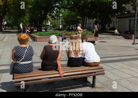 Rear view of people sitting on a bench watching two young musicians busking in Gedimino Prospektas in Vilnius, Lithuania. Stock Photo