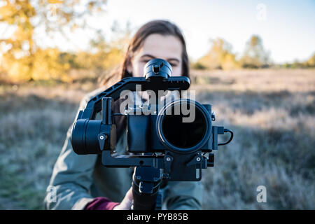 Female videographer holding a gimbal with mirrorless camera. Woman with stabilized camera rig filming outdoors on a sunny afternoon Stock Photo