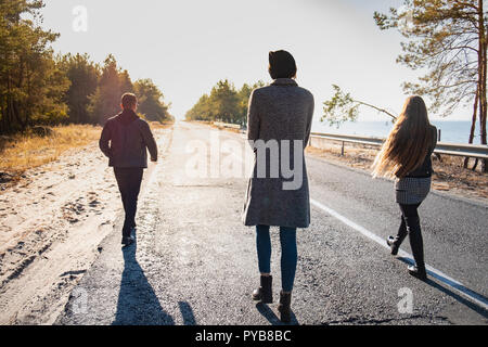 Group of people walk along the road at the seaside. Three young persons walking in beautiful nature scene in late summer or autumn Stock Photo