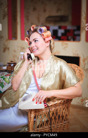 Cute woman in bathrobe and hair curlers talking on the phone while sitting in the kitchen. Retro and vintage. Stock Photo