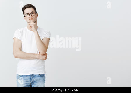 Thoughtful serious-looking male coworker thinkings, trying make up idea, touching chin raising one eyebrow in interest making assumptions or choice, stuck with decision over gray background Stock Photo