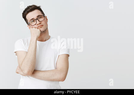 What boring day at work. Portrait of indifferent apathic and unintersted attractive young man in glasses leaning head on hand from boredom looking at camera with no interest and joy over grey wall Stock Photo