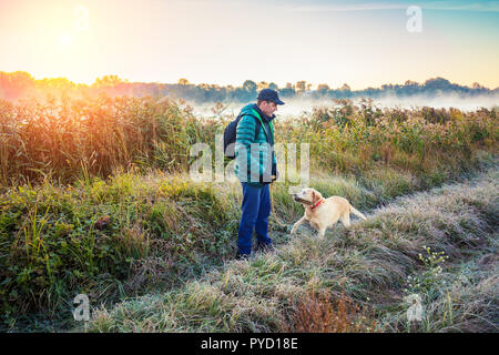 A man with Labrador retriever dog walking on the rural road near the lake in the early autumn morning Stock Photo