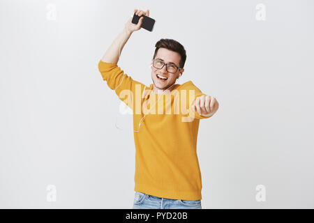 Guy enjoying autumn with favorite tracks in earphones dancing joyfully pointing at camera and raising hand as holding smartphone smiling broadly lifting mood of listening music in earphones from app Stock Photo