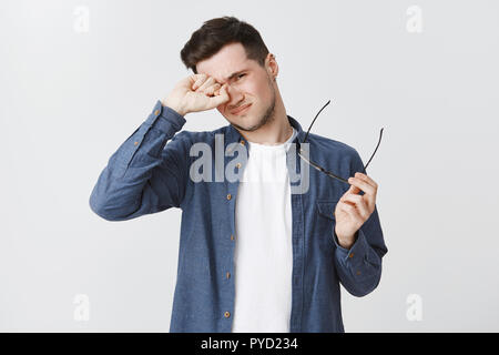 Waist-up shot of tired male freelancer taking of glasses as working long near computer on gig economy project rubbing eye and frowning being exhausted standing drained and upset over grey wall Stock Photo