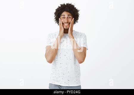 Face aches from constant smile, man got in love. Cute carefree and happy hispanic male with tanned skin and afro hairstyle, holding palms on cheeks and smiling broadly from joy and amusement Stock Photo