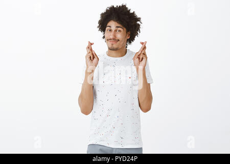 Lucky man crossing fingers, making sure he will win, hoping for bet to play. Portrait of charming relaxed hipsanic guy with moustache and afro hairstyle, smirking and gazing at camera with confidence Stock Photo