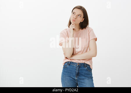 Girl wonders if she can afford one more t-shirt. Determined creative and cute woman, standing in thoughtful pose, thinking while folding lips and looking at upper left corner curiously, touching chin Stock Photo