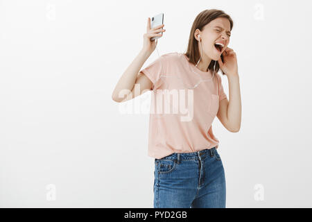Girl feeling happy and great, singing along and enjoying nice bits of new earphones, yelling while listening favorite song, smiling broadly, holding smartphone in raised arm, dancing over grey wall Stock Photo
