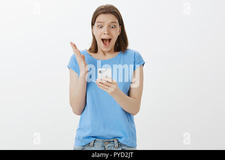 Portrait of cute good-looking european woman in blue t-shirt shouting from excitement and shaking hand while staring surprised and amazed at smartphone screen, being impressed over grey wall Stock Photo
