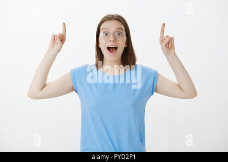 Positive excited european girlfriend in blue t-shirt, raising hands and pointing upwards with forefinger, saying yeah and smiling broadly, being impressed and fascinated with great copy space Stock Photo
