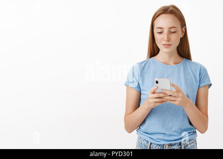 Cute urban redhead female student answering message while driving in bus texting via smartphone looking happy and relaxed at device screen standing in casual blue t-shirt over gray background Stock Photo
