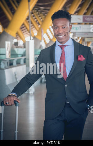 Close uo of one young and attractive black businessman is waiting for a car outside of the airport wearing a black suit with a red tie and carrying a  Stock Photo