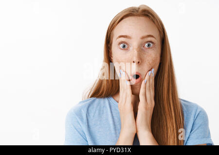 Headshot of shocked and surprised gorgeous redhead female with freckles folding lips and gasping from amazement and shook holding hands on face standing impressed over gray background Stock Photo