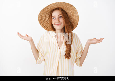 No big deal. Portrait of carefree optimistic charming kind female with ginger hair and freckles in cute straw hat and yellow blouse making shoulder shrugg with hands spread and innocent look Stock Photo