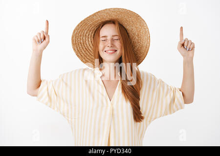 Waist-up shot of optimistic dreamy and happy girlfriend with red hair and freckles closing eyes with joyful look pointing up with raised hands wearing cute summer straw hat and striped yellow blouse Stock Photo