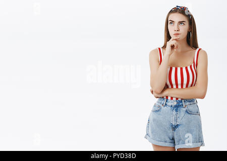 Creative girlfriend trying think up plan of reading boyfriend messages standing in trendy pinup outfit with headband and striped red top making hmm look with hand on chin, looking upwards and thinking Stock Photo