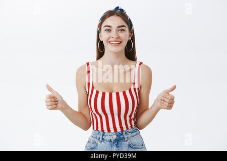 Satisfied beautiful slim woman in striped pin-up style top and headband smiling broadly showing thumbs up expressing like and approval being supportive giving positive feedback about product Stock Photo
