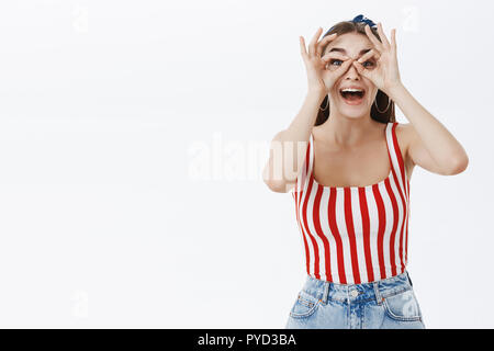 Let us have fun. Funny enthusiastic attractive young woman in striped top and shorts making goggles with fingers over eyes and looking through at camera joyfully smiling broadly from amusement Stock Photo