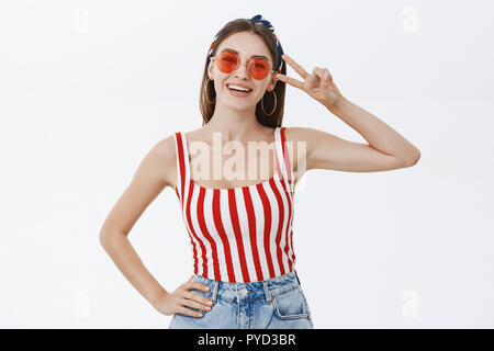 Indoor shot of stylish emotive young attractive pinup woman in striped top and trendy round sunglasses showing peace or victory sign near face, smiling joyfully holding hand on waist over white wall Stock Photo