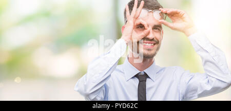 Young business man with happy face smiling doing ok sign with hand on eye looking through fingers Stock Photo