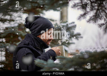 Side view of attractive guy drinking fresh hot beverage while standing near conifer trees on beautiful winter day Stock Photo