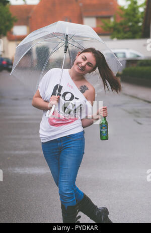 A young woman with an umbrella in her hand dances in the rain holding a bottle of Heineken non-alcoholic beer Stock Photo