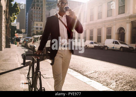 Businessman going to work by bicycle. African man walking with cycle and drinking coffee on city street. Stock Photo