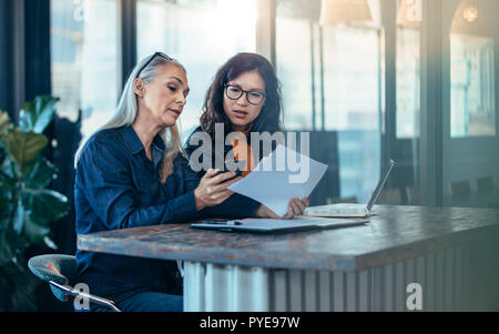 Two business woman sitting at a table and working together. Senior executive showing her phone and papers to young colleague at office. Stock Photo