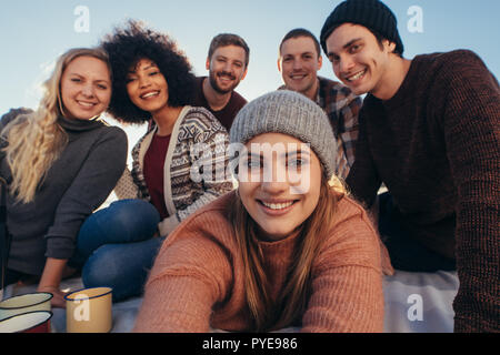 Beautiful young woman taking selfie with friends sitting around at the beach. Group of friends taking selfie on the seashore. Stock Photo