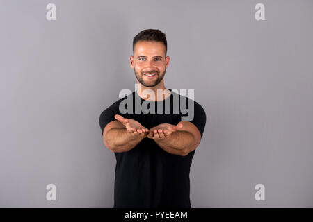 A handsome young man in a black tshirt showing something with his palms in front of a grey background in the studio. Stock Photo