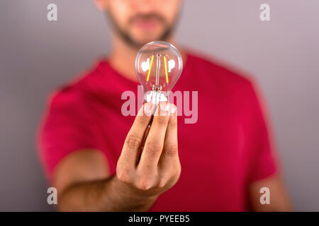 A young man in a red tshirt holding a lightbulb and standing in front of a grey background in the studio. Stock Photo
