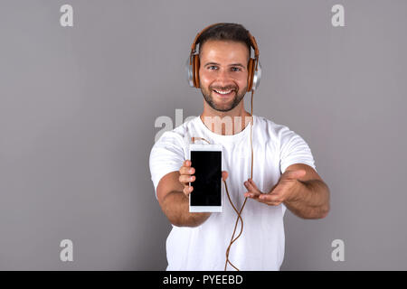 A handsome young man in a white tshirt listening to music on his headphones and showing his phone to the camera while standing in front of a grey back Stock Photo