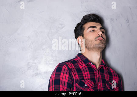 A serious handsome young man in a red checkered shirt standing in front of a grey wall in a studio. Stock Photo