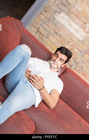 A serious handsome young man in a white tshirt laying on a sofa and using his smartphone. Stock Photo