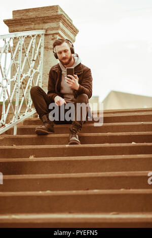 A handsome young hipster man sitting on the stairs and taking a selfie with his smartphone. Stock Photo