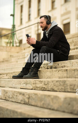A handsome young hipster man sitting on the stairs and checking his smartphone. Stock Photo