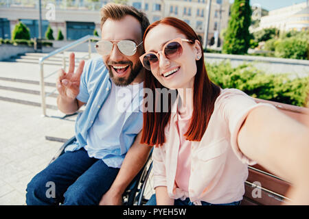 camera point of view of happy boyfriend in wheelchair and attractive girlfriend looking at camera in city, man showing peace sign Stock Photo