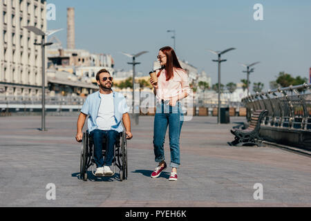 handsome boyfriend in wheelchair and girlfriend with disposable coffee cup having walk on street Stock Photo