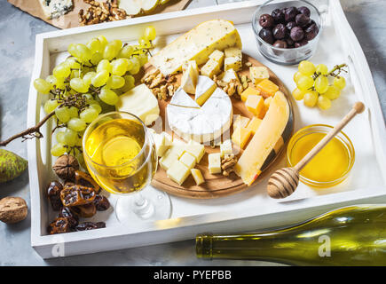 Different types of cheese on wooden board, olive, fruits, almond and wine glasses on white tray on concrete table Stock Photo